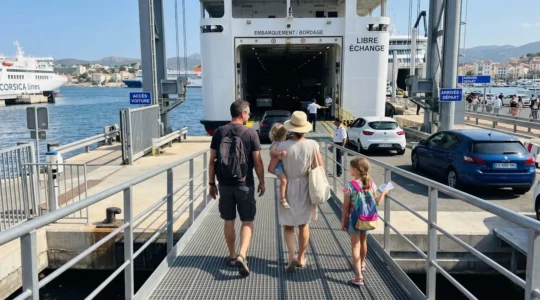 Une famille de quatre personnes vue de dos monte la passerelle d'embarquement d'un ferry moderne, leur voiture visible en arrière-plan sous un ciel méditerranéen ensoleillé