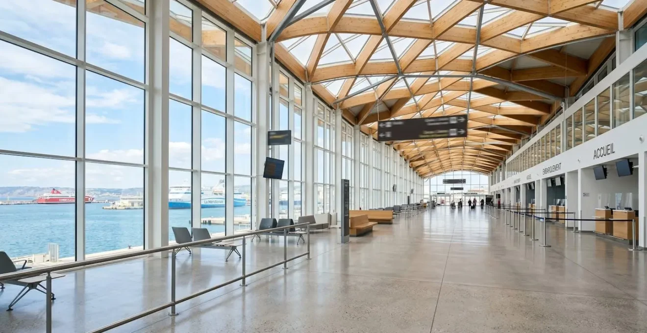 Vue large de l'intérieur d'un terminal de ferry méditerranéen contemporain, grandes baies vitrées laissant entrer la lumière naturelle, architecture moderne épurée, hall spacieux vide, panneaux directionnels complètement floutés