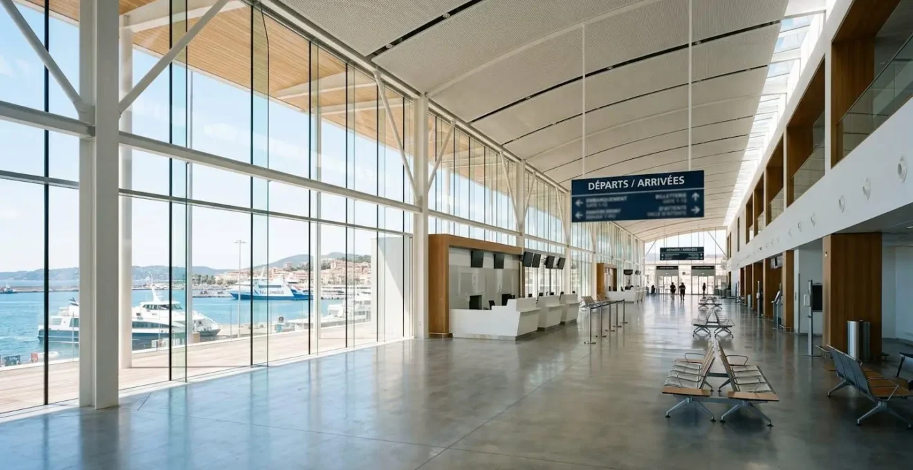 Vue large de l'intérieur d'un terminal de ferry méditerranéen contemporain, grandes baies vitrées laissant entrer la lumière naturelle, architecture moderne épurée, hall spacieux vide, panneaux directionnels complètement floutés